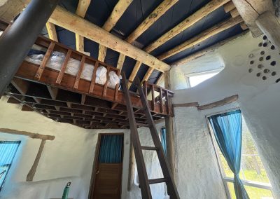 Wooden loft ladder leading to sleeping area with exposed beams and storage in hand-built earth building at Travel Magick Spiritual Retreat Center