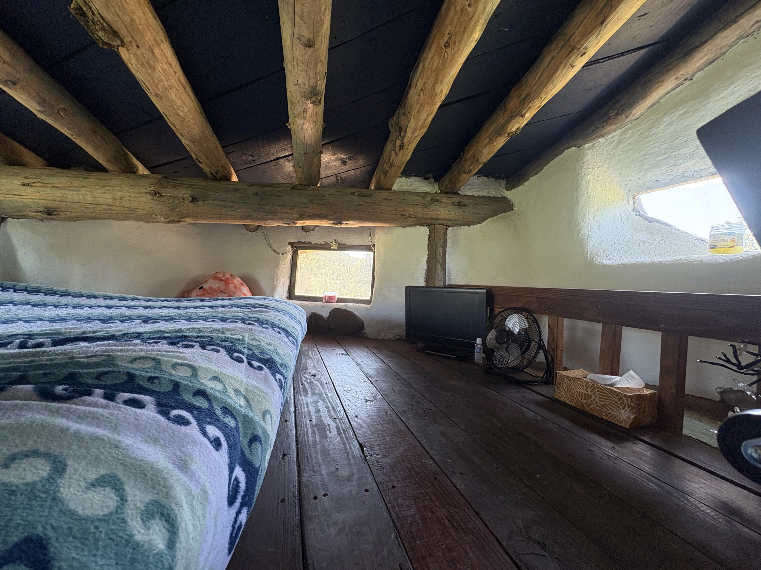 Cozy loft bedroom with exposed wooden beams and natural light windows in hand-built earth building at Travel Magick Spiritual Retreat Center